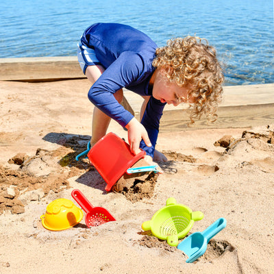 A young child with curly hair plays in the sand by a lake, using a red bucket with the HABA 5 Piece Basic Sand Toys Set  scattered nearby.