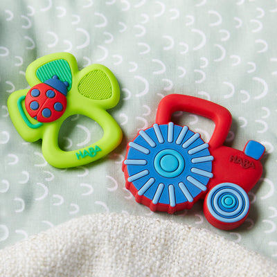 Two colorful baby teether toys on a patterned fabric background, one shaped like a clover with a ladybug and the other like a tractor.