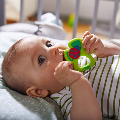 A baby holding a green Shamrock Silicone Teether toy with a ladybug design, lying on a patterned pillow.