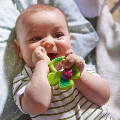 A baby chewing their fist holding a green Shamrock Silicone Teether toy with a ladybug design, lying on a patterned pillow.