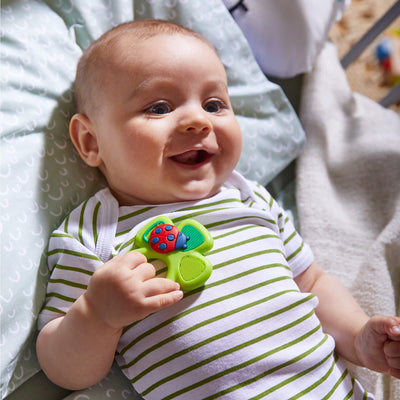 A baby laying down smiling holding a green Shamrock Silicone Teether toy with a ladybug design, lying on a patterned pillow.