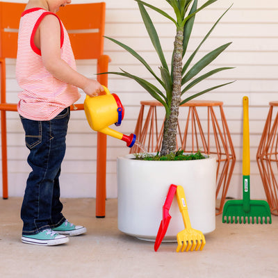 Child watering a potted plant using a Spielstabil yellow watering can with toy gardening tools nearby.