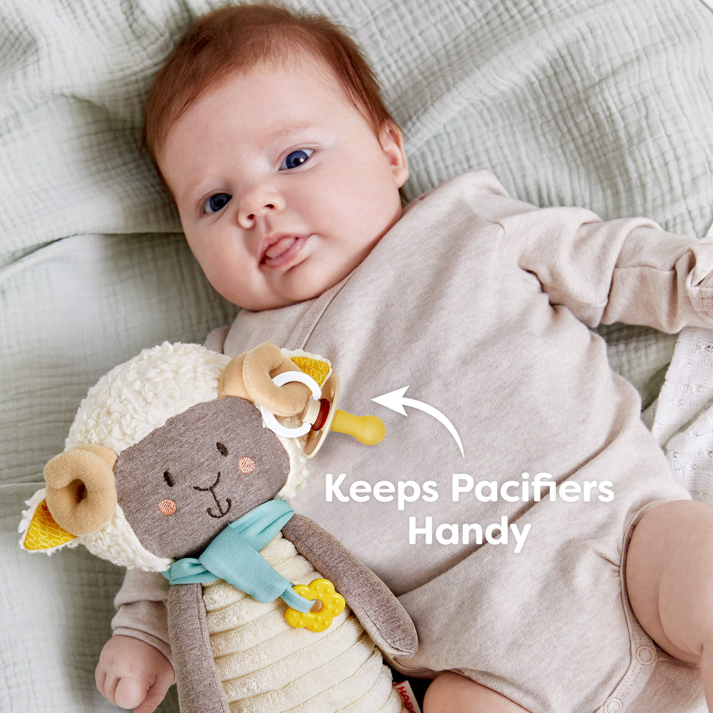 A baby with a plush sheep toy and pacifier on a soft blanket with the text "Keeps Pacifiers Handy"