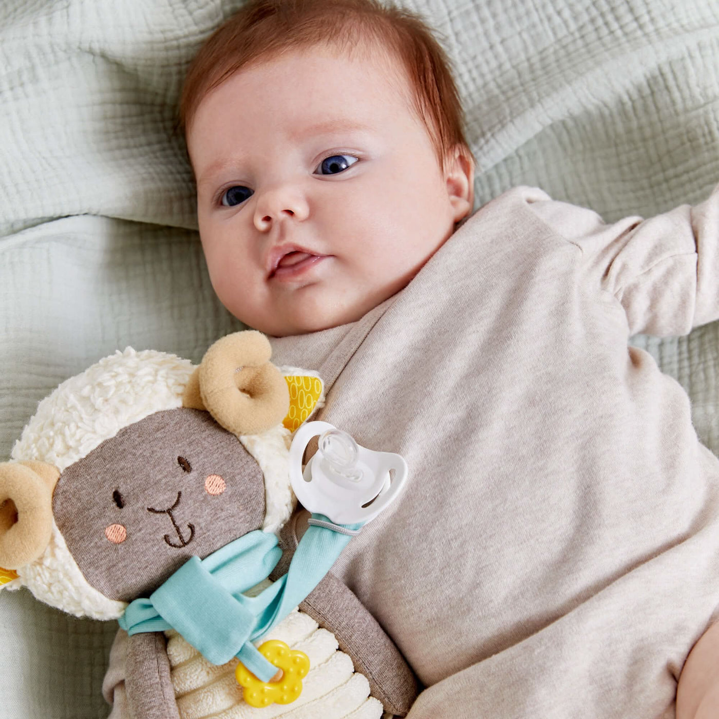 A baby lying on a quilted blanket with a ram plush toy with an attached pacifier.